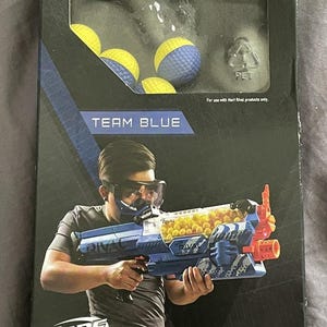 May include: A box of Nerf Rival high-impact rounds. The box is black with blue and white accents and features the text "TEAM BLUE" and "50x HIGH-IMPACT ROUNDS". The box also shows a person holding a blue and orange Nerf Rival blaster.