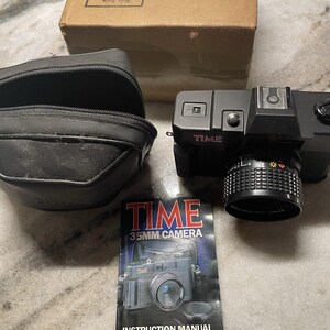 Puede incluir: Una cámara TIME 35mm negra con una lente, una funda de transporte negra y un manual de instrucciones. El manual de instrucciones dice "TIME 35MM CAMERA" y "INSTRUCTION MANUAL". También hay una caja de cartón.
