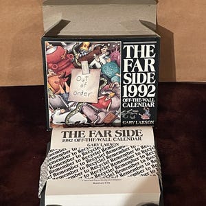 Op de afbeelding: Een "The Far Side" off-the-wall kalender uit 1992 in de originele doos. De doos heeft een cartoon illustratie en de tekst "Out of order". De kalender is open en toont de dagelijkse pagina's en een zwarte basis.