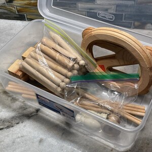 Puede incluir: Un recipiente de almacenamiento de plástico transparente lleno de piezas de juguetes de madera. El recipiente contiene clavijas de madera, bloques y una rueda de madera. Algunas piezas están en bolsas de plástico. El recipiente está sobre una superficie de mármol.