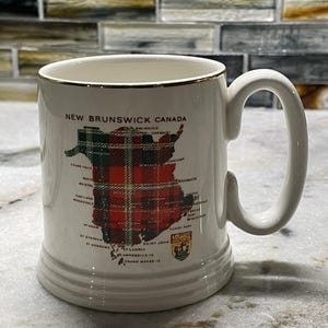 May include: White ceramic mug with a gold rim, displaying a map of New Brunswick, Canada, with a red and green tartan pattern. The mug has a large handle and is labelled with city names.