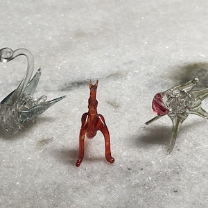 May include: Three glass figurines: a swan with a clear body and blue wings, a red and orange abstract figure, and a clear figure with a pink head and eight legs.