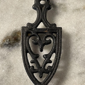 May include: Black cast iron trivet with a decorative heart design. The trivet has a small loop at the top for hanging.