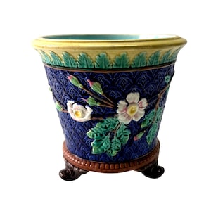 May include: A decorative blue ceramic planter with a textured pattern and floral relief. The planter features white and pink flowers with green leaves, a yellow rim, and a brown base with three feet. A unique piece for home decor.