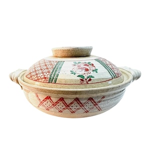 May include: A beige ceramic Japanese donabe pot with a lid. The lid features a floral design with red and green accents, along with a geometric pattern. The pot has two handles and a matching lid knob.