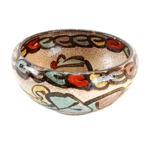 Puede incluir: Un cuenco de cerámica pequeño y redondo con interior color crema y borde marrón oscuro. El cuenco está decorado con coloridos patrones florales y geométricos en rojo, azul, verde y marrón. El cuenco tiene un aspecto agrietado y envejecido.