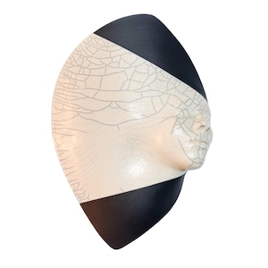May include: A wall-mounted ceramic sculpture shaped like a stylized human face in profile. The piece features a matte black finish on the top and bottom sections, with a central cream-colored area displaying a distinct, dark crackle glaze pattern.