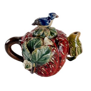 May include: A ceramic teapot shaped like a large red strawberry with a dark brown handle and a green spout. The lid features a small blue bird perched on green leaves. The body of the teapot is decorated with sculpted green leaves and white flowers.