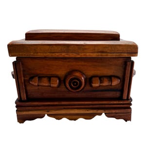 May include: A rectangular wooden box with a rich brown finish, featuring a tiered lid, a central circular carved detail, and two horizontal carved accents on the front panel. The base has a scalloped edge design with vertical side supports.