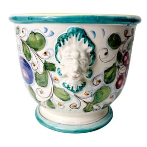 May include: A white ceramic planter with a turquoise rim and base. The planter features a raised relief of a face and is decorated with green leaves, red berries, and blue and yellow accents. The planter is a decorative item.