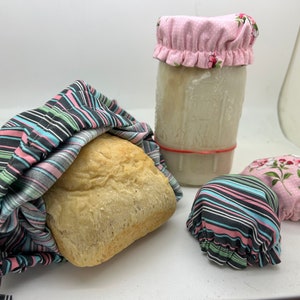May include: Three reusable food covers made of fabric with a striped pattern in black, pink, and blue. One cover is over a loaf of bread, another is over a glass jar, and the third is on a table.