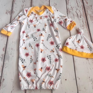 May include: A white baby gown with a floral and bee print. The gown has yellow trim and comes with a matching hat.