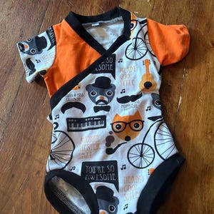 May include: A baby bodysuit with a wrap-style front. The top is orange with black trim, and the body features a white background with black illustrations of mustaches, musical instruments, and the text "You're so awesome".