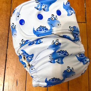 Op de afbeelding: Een witte stoffen luier met een herhaald patroon van blauwe cartoon dinosaurussen. De luier heeft blauwe drukknoopsluitingen en elastische randen. De dinosaurussen hebben vriendelijke uitdrukkingen op een witte achtergrond.