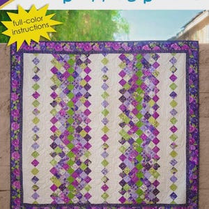 May include: A quilt pattern named "Stepping Stones" by Cozy Quilt Designs. The quilt design has a white backdrop with vertical columns of purple, green, and white diamond patterns. The border features a floral design. The pattern includes full-colour instructions and is by Daniela Stout.