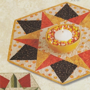 May include: A sewing pattern cover titled Atkinson Designs one of life's simple pleasures ATK-147. It features a hexagonal quilted table topper with black, orange, and yellow star patterns, and a bowl of candy corn with a white candle.