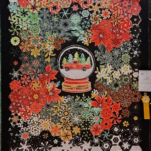 May include: A black and white quilt with a red, green, and white winter scene in the center. The scene features a snow globe with a red truck inside. The quilt is made up of various geometric shapes and patterns.