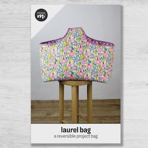 May include: A quilted fabric project bag featuring a floral pattern with pink flowers, green vines, and blue accents on a white background. The bag has a distinct curved top edge and purple trim. The text "laurel bag a reversible project bag" is below.