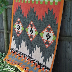 May include: A quilt pattern called "Montana" by Sherri Noel. The quilt is made with a variety of fabrics in black, white, orange, red, and green. The quilt is hanging on a wooden fence.