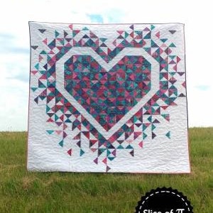 May include: A white quilt featuring a large pink and blue heart at its centre. The heart is composed of smaller triangles and is surrounded by a burst of smaller triangles. The quilt is titled "Exploding Heart" and is made by Slice of Pi Quilts.