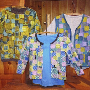 May include: Three colorful patchwork jackets and a blue shirt hang on wooden hangers. The jackets feature a variety of square patterns in shades of yellow, blue, green, and purple. The jackets are displayed against a wooden background.