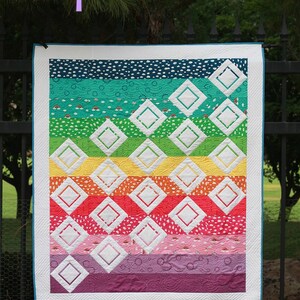 May include: A rainbow-colored quilt with white diamond shapes. The quilt is hanging on a black metal fence. The quilt is titled "Sprinkles" and is a beginner foundation paper pieced pattern. The pattern includes instructions for baby, throw, and twin sizes.