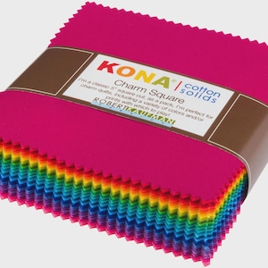 May include: A stack of square fabric pieces with pinked edges, wrapped in a brown paper band labeled KONA cotton solids Charm Square. The fabric layers display a spectrum of colors including pink, orange, yellow, green, blue, and purple.