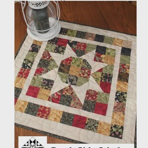 May include: A square quilt topper titled Sunny Days measuring 25 inches by 25 inches by Creek Side Stitches #323. The quilt features a central star pattern with a multi-colored patchwork design in shades of red, green, yellow, and black.