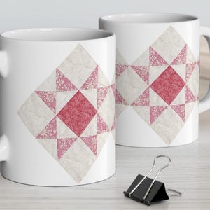 May include: Two white ceramic mugs with a quilt-inspired design. Each mug features a central red square surrounded by white and pink geometric shapes. The mugs are on a light-colored surface.