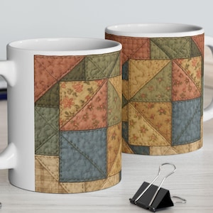 May include: Two white ceramic mugs with a patchwork quilt design. The design features squares and triangles in shades of peach, green, blue, and tan, with floral and solid patterns. The mugs are on a light-coloured wooden surface.
