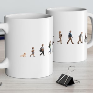 Può includere: Due tazze in ceramica bianca con una grafica che raffigura le fasi della vita umana. Il disegno mostra un bambino che gattona, seguito da bambini, adulti e una persona anziana in sedia a rotelle. Le tazze sono su una superficie chiara.