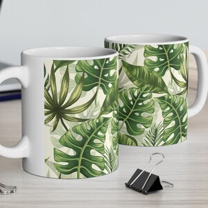 May include: Two white ceramic mugs featuring a tropical leaf design. The mugs have a white background with green leaves of various shapes and sizes.