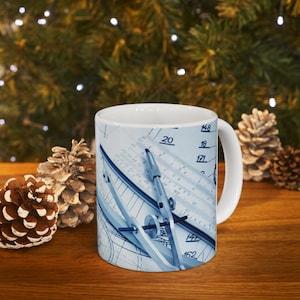 Pode incluir: Uma caneca de cerâmica branca com um desenho azul e branco de um compasso e uma régua em um projeto. A caneca tem uma alça do lado direito.