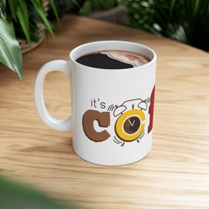 Puede incluir: Taza de café blanca con un gráfico marrón y amarillo que dice "it's coffee time" con un despertador de dibujos animados. La taza está llena de café negro.