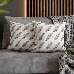 Pode incluir: Duas almofadas quadradas com fundo branco apresentando um padrão repetitivo de pequenos cachos de rosas cor-de-rosa com folhas e caules verdes, dispostos em filas diagonais na superfície de tecido de cada almofada.