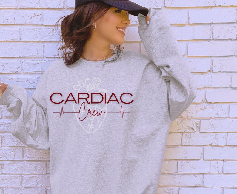 Puede incluir: Una sudadera gris con el texto "CARDIAC Crew" en letras color granate y un gr&aacute;fico de coraz&oacute;n estilizado. El gr&aacute;fico del coraz&oacute;n es blanco con un contorno granate y una l&iacute;nea de latido del coraz&oacute;n por encima y por debajo.
