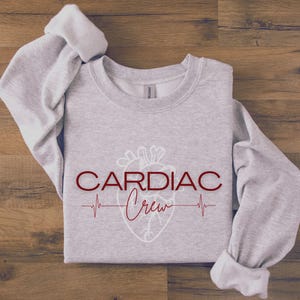 Puede incluir: Sudadera gris con un contorno blanco de un coraz&oacute;n y el texto "CARDIAC Crew" en rojo con una l&iacute;nea de latido del coraz&oacute;n debajo.
