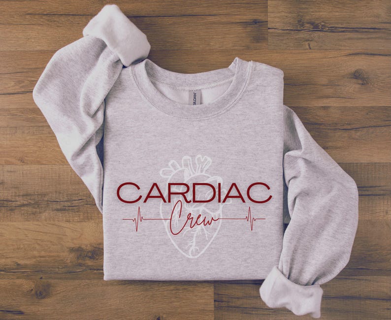 Puede incluir: Sudadera gris con un contorno blanco de un coraz&oacute;n y el texto "CARDIAC Crew" en rojo con una l&iacute;nea de latido del coraz&oacute;n debajo.