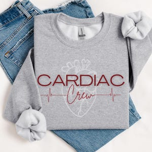 Puede incluir: Sudadera gris con un coraz&oacute;n blanco y el texto "CARDIAC Crew" en granate. Una l&iacute;nea roja de ECG est&aacute; debajo del texto.