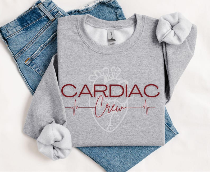 Puede incluir: Sudadera gris con un coraz&oacute;n blanco y el texto "CARDIAC Crew" en granate. Una l&iacute;nea roja de ECG est&aacute; debajo del texto.