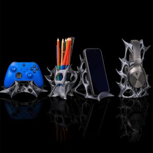 May include: A set of silver-coloured, abstract-design holders for tech and office items. Includes a blue game controller, a pencil holder with coloured pencils, a smartphone, and headphones. All items are displayed against a black background.