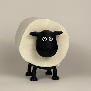 Puede incluir: Un porta rollo de papel higi&eacute;nico con forma de oveja en blanco y negro. La oveja tiene la cara negra con ojos blancos y nariz negra. El rollo de papel higi&eacute;nico es blanco y se mantiene en su lugar por el cuerpo de la oveja.