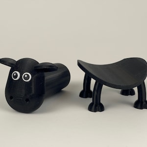 Puede incluir: Dos figuritas negras impresas en 3D. Una es una oveja con ojos blancos y la otra es un peque&ntilde;o taburete con cuatro patas.