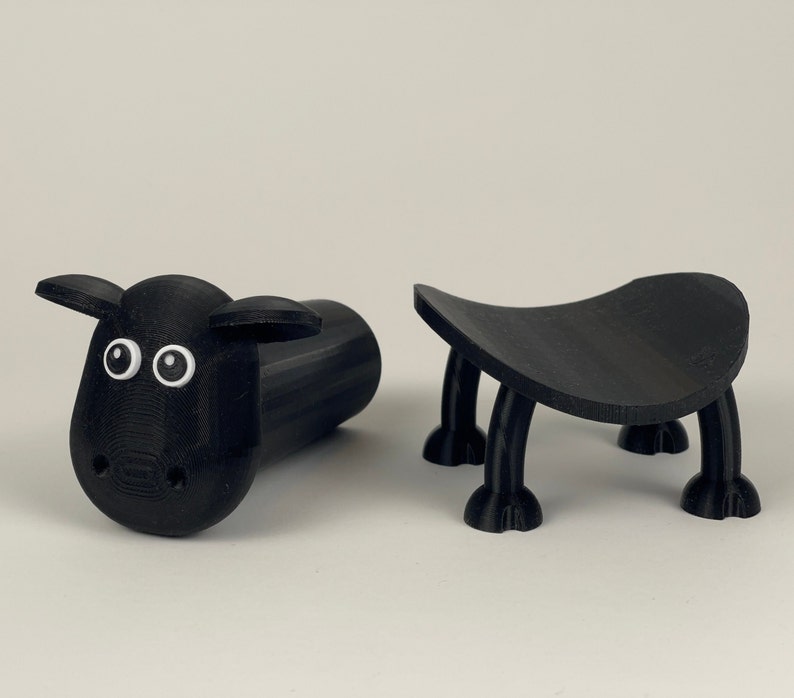 Puede incluir: Dos figuritas negras impresas en 3D. Una es una oveja con ojos blancos y la otra es un peque&ntilde;o taburete con cuatro patas.