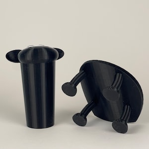 Puede incluir: Dos objetos negros impresos en 3D. Uno es un objeto alto y cil&iacute;ndrico con dos peque&ntilde;as orejas redondeadas en la parte superior. El otro es un objeto plano y circular con cuatro patas.