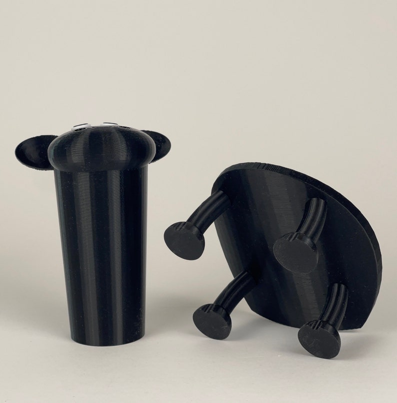 Puede incluir: Dos objetos negros impresos en 3D. Uno es un objeto alto y cil&iacute;ndrico con dos peque&ntilde;as orejas redondeadas en la parte superior. El otro es un objeto plano y circular con cuatro patas.