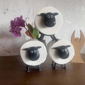 Puede incluir: Tres porta rollos de papel higi&eacute;nico blancos con forma de oveja con caras y patas negras. Las ovejas est&aacute;n apiladas una encima de la otra.