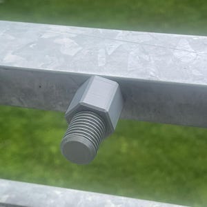 Op de afbeelding: Close-up van een grijze bout en moerconstructie bevestigd aan een metalen balk. De bout heeft een schroefdraadas en een zeskantige kop. De metalen balk is zilverkleurig, met een vage groene achtergrond.
