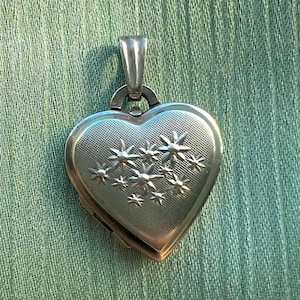 May include: A silver-toned heart-shaped locket pendant featuring a textured surface with a cluster of embossed star-shaped floral motifs on the front. The locket includes a polished metal bail at the top for attachment to a chain or necklace.
