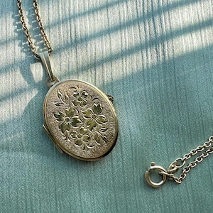May include: An oval gold-tone locket pendant featuring an embossed floral pattern on the front surface. The locket is attached to a delicate gold-tone chain that includes a circular spring ring clasp at the end for secure fastening.